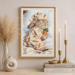 Peut inclure: Une impression d'art encadr&eacute;e repr&eacute;sentant un lapin endormi portant un pyjama &agrave; motif de carottes et un bonnet de nuit assorti. Le lapin tient une grosse carotte orange tout en se reposant sur un oreiller blanc. L'illustration utilise des tons pastel.