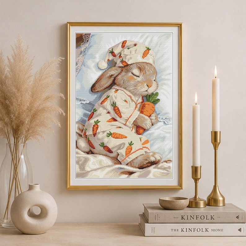 Peut inclure: Une impression d'art encadr&eacute;e repr&eacute;sentant un lapin endormi portant un pyjama &agrave; motif de carottes et un bonnet de nuit assorti. Le lapin tient une grosse carotte orange tout en se reposant sur un oreiller blanc. L'illustration utilise des tons pastel.