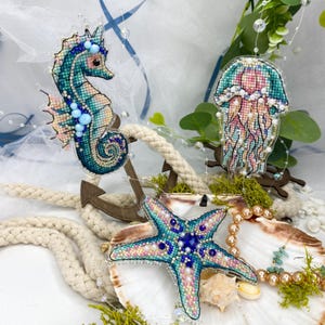 May include: Embroidered sea creature ornaments, including a seahorse, jellyfish, and starfish, in shades of blue, pink, and green. The ornaments are decorated with beads and pearls, evoking a nautical theme. They are displayed with rope, shells, and greenery.