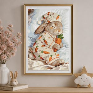 Peut inclure: Une broderie au point de croix encadr&eacute;e repr&eacute;sentant un lapin endormi portant un pyjama et un bonnet de nuit, tous deux orn&eacute;s de motifs de carottes orange. Le lapin repose sur un oreiller blanc et tient une grosse carotte orange.