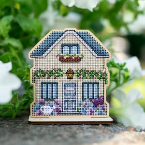 Puede incluir: Una casa de madera bordada a punto de cruz con tejado azul, ventanas y puerta. La casa está decorada con detalles florales, una pequeña regadera y una mariposa morada. La casa está situada sobre un fondo de follaje verde y flores blancas.