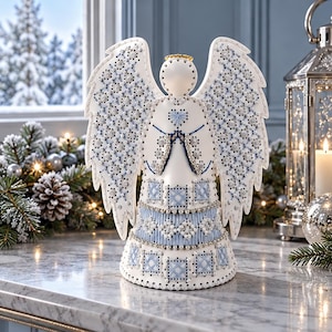 May include: A white angel figurine featuring intricate blue and silver thread embroidery on its wings and gown. The design includes geometric patterns, a small gold halo, and beaded accents, creating a detailed, textured appearance.