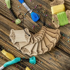 Pode incluir: Um conjunto de 10 porta-bobinas de madeira com um design floral decorativo. As bobinas são dispostas em forma de leque e são mantidas juntas por um cordão preto. As bobinas são feitas de madeira natural e têm uma cor castanho claro.