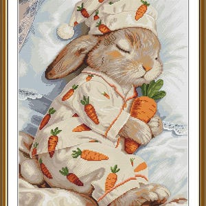 Peut inclure: Un motif de broderie au point de croix repr&eacute;sentant un lapin brun endormi portant un pyjama cr&egrave;me et un bonnet de nuit assorti, tous deux orn&eacute;s d'un motif de carottes orange. Le lapin serre une grosse carotte orange contre sa poitrine.