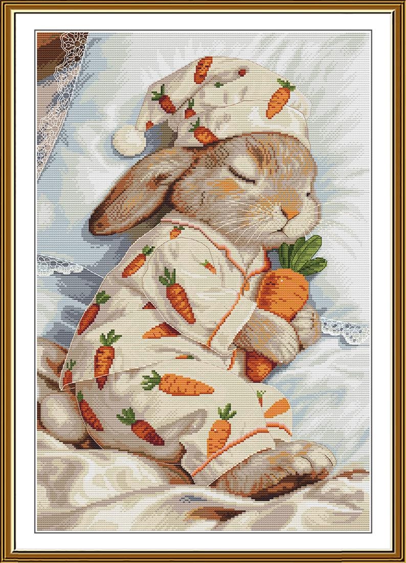 Peut inclure: Un motif de broderie au point de croix repr&eacute;sentant un lapin brun endormi portant un pyjama cr&egrave;me et un bonnet de nuit assorti, tous deux orn&eacute;s d'un motif de carottes orange. Le lapin serre une grosse carotte orange contre sa poitrine.