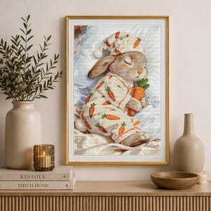 Peut inclure: Une broderie au point de croix encadr&eacute;e repr&eacute;sentant un lapin endormi portant un bonnet de nuit et un pyjama assortis, tous deux orn&eacute;s d'un motif de carottes orange r&eacute;p&eacute;titif. Le lapin repose sur un oreiller blanc en tenant une grosse carotte.