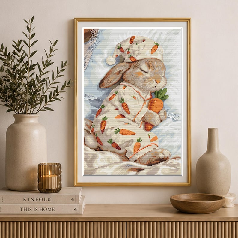 Peut inclure: Une broderie au point de croix encadr&eacute;e repr&eacute;sentant un lapin endormi portant un bonnet de nuit et un pyjama assortis, tous deux orn&eacute;s d'un motif de carottes orange r&eacute;p&eacute;titif. Le lapin repose sur un oreiller blanc en tenant une grosse carotte.