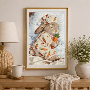 Peut inclure: Une impression d'art encadr&eacute;e repr&eacute;sentant un lapin endormi portant un pyjama et un bonnet de nuit assorti, tous deux d&eacute;cor&eacute;s d'un motif de carottes. Le lapin repose sur un oreiller blanc et tient une carotte orange dans ses pattes.