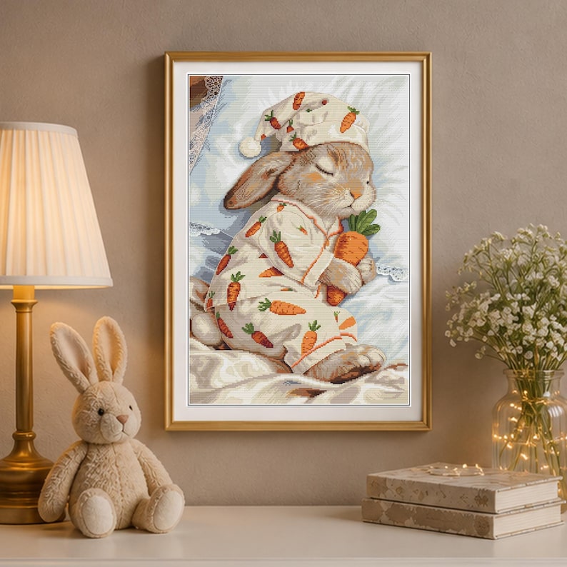 Peut inclure: Une broderie au point de croix encadr&eacute;e repr&eacute;sentant un lapin brun clair dormant sur un oreiller blanc. Le lapin porte un bonnet de nuit et un pyjama assortis avec un motif de carottes orange et tient une grosse carotte orange dans ses pattes.