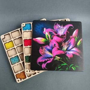 Könnte beinhalten: Holz-Bastel-Organizer mit mehreren Fächern, gefüllt mit bunten Perlen. Ein dekoratives Holzquadrat mit einem lebendigen Blumendesign mit rosa und lila Lilien ist ebenfalls vorhanden. Die Artikel sind auf einer grauen Oberfläche angeordnet.