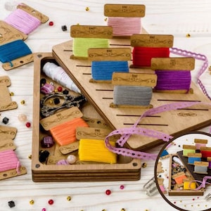 May include: Wooden box with a hinged lid, containing compartments for storing thread spools. The box has a built-in thread cutter and a small compartment for storing other sewing supplies. The box is open and the compartments are filled with colourful thread spools.