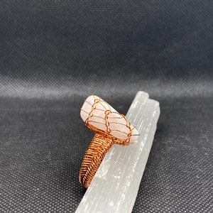 May include: A pink rose quartz stone wrapped in copper wire, forming a ring. The ring is displayed on a white selenite charging plate.