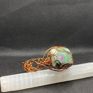 May include: A copper wire wrapped bracelet featuring a green and pink gemstone. The bracelet is displayed on a white selenite charging plate.