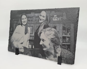 Può includere: Una foto in bianco e nero di tre donne è stampata su una placca di ardesia. Le donne sorridono e sembrano essere in un ambiente da cucina.