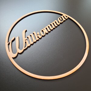 Könnte beinhalten: Ein Holzschild mit dem deutschen Wort "Willkommen" (Willkommen) in einer kursiven Schriftart. Das Wort ist diagonal über der Mitte eines kreisförmigen Holzrahmens angeordnet. Das Schild ist aus hellem Holz gefertigt und vor einem dunklen Hintergrund platziert.
