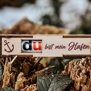 Pu&ograve; includere: Una scatola di legno con un cassetto scorrevole che dice "du bist mein Hafen" (tu sei il mio porto) in tedesco. La scatola ha una piccola ancora intagliata sul lato sinistro e un'etichetta rossa, bianca e nera con la parola "du" (tu) nel cassetto.