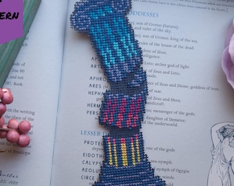 May include: A handmade cross-stitch bookmark in a column design, featuring shades of blue, pink, and yellow. The bookmark is placed on an open book with text and a purple flower in the background. The words "PDF PATTERN" are in a purple banner.