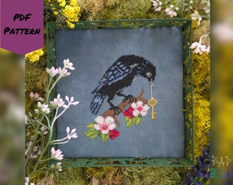 May include: Cross-stitch pattern featuring a black crow holding a golden key, perched on a flowering branch. The design is set against a blue background within a green frame. The words "PDF PATTERN" are in the upper left corner.
