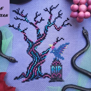 May include: Cross-stitch pattern showing a spooky tree with pink and blue details, a tombstone, and a blue bird. The text "PDF PATTERN" is in the upper left corner. Two black snakes are on the sides. The text "BAD Stitch" is in the lower right corner.