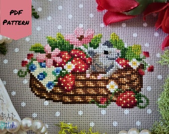 May include: Cross stitch pattern featuring a gray mouse sitting in a brown basket filled with red strawberries, pink and blue flowers, and green leaves. The pattern is on a gray background with white polka dots. The text "PDF PATTERN" is in a pink box at the top left of the image.