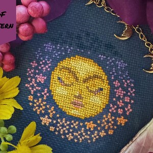 May include: Cross-stitch pattern featuring a yellow moon with closed eyes, surrounded by a ring of pink and purple stars, on a dark blue fabric. The words "PDF PATTERN" are in a purple banner. A gold chain with moon charms is visible.