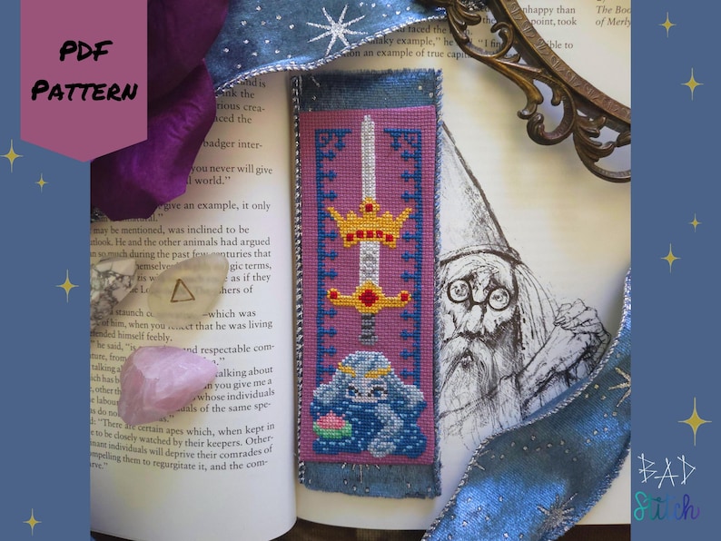 May include: A cross-stitch bookmark with a sword and crown design, featuring a blue bunny with a flower. The bookmark is on a book with a wizard illustration and the text "PDF PATTERN". The bookmark is approximately 20 cm long.