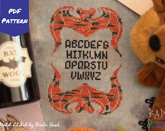 May include: A cross-stitch pattern featuring a bat design with the alphabet in black letters on a white background. The pattern is stitched with orange and black thread. The text "PDF PATTERN" is in blue letters on a white background. The text "BAT WOOL" is in black letters on a white background. The text "ABCDEFG HIJKLMN OPQRSTU VWXYZ" is in black letters on a white background. The text "Model stitched by Bronte Count" is in black letters on a white background. The text "BAD Stitch" is in black letters on a white background.