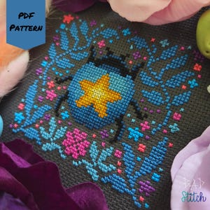 May include: Cross-stitch pattern featuring a stylized beetle with a yellow star on a blue body, surrounded by blue leaves and pink and blue flowers on a dark gray background. The text "PDF PATTERN" is in a blue box.