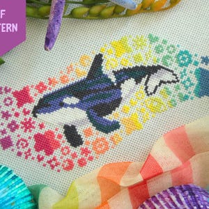 May include: A cross-stitch pattern on white fabric featuring a black and white orca whale surrounded by a trail of colorful stars, moons, and sparkles in a rainbow gradient. The text PDF PATTERN is visible in a purple banner in the top left corner.