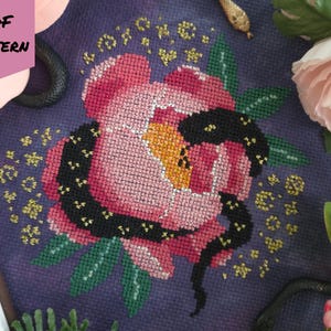 May include: A cross-stitch pattern on dark purple fabric featuring a large pink peony flower with a black snake coiled around it, surrounded by small yellow stars and dots. The image includes a pink tag with the text PDF PATTERN.