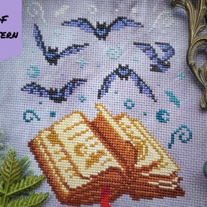 May include: A cross stitch pattern featuring a book with bats flying around it. The book is open and has a red ribbon bookmark. The bats are blue and purple and are flying in a circle around the book. The background is a light purple color.
