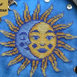 May include: A cross-stitch embroidery pattern of a sun and moon with a blue background. The sun is yellow and has a face with a smile. The moon is blue and has a crescent shape. The pattern is surrounded by a border of small, white beads.