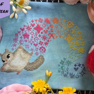 May include: A cross-stitch pattern on light blue fabric featuring a flying squirrel with a trail of colorful, sparkling stars and hearts in a rainbow gradient. The text PDF PATTERN is in the top left corner and BAD Stitch is in the bottom right.