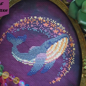 May include: Cross-stitch design of a whale swimming through a starry arc. The whale is blue and white, with an orange and white belly. The background is purple. The text "PDF PATTERN" is in the upper left corner.