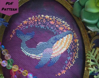May include: Cross-stitch pattern featuring a whale swimming through a starry arc. The whale is blue and white, with an orange and white belly. The background is purple. The text "PDF PATTERN" is in the upper left corner.