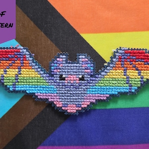May include: A cross-stitch bat with rainbow wings and a purple body, set against a colorful striped background. The bat has small black eyes and pink accents. The words "PDF PATTERN" are in a purple box in the upper left corner.