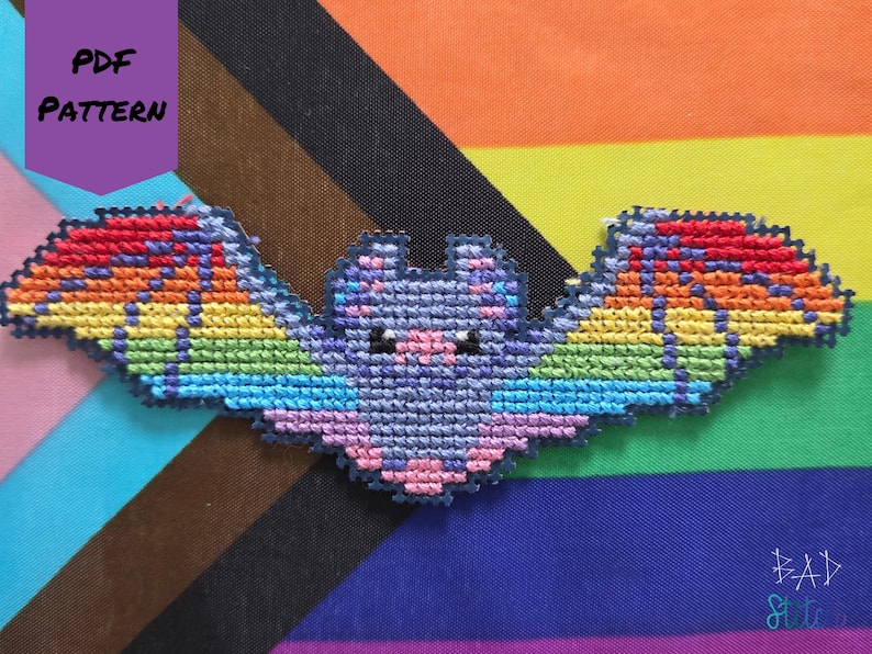 May include: A cross-stitch bat with rainbow wings and a purple body, set against a colorful striped background. The bat has small black eyes and pink accents. The words "PDF PATTERN" are in a purple box in the upper left corner.