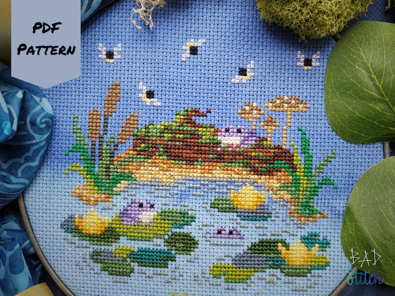 Op de afbeelding: Borduurpatroon met een fantasierijke sc&egrave;ne van kikkers op waterlelies in een vijver. Het patroon bevat een blauwe lucht, groene waterlelies, gele bloemen en bruine rietstengels. Het patroon is in de linkerbovenhoek getiteld "PDF PATTERN".