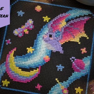 May include: A cross-stitch pattern featuring a dragon on a crescent moon, surrounded by stars and a small planet. The design uses a palette of blues, purples, pinks, and yellows against a black background. The words "PDF PATTERN" are visible.