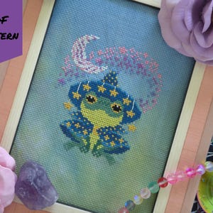 May include: Cross-stitch pattern of a whimsical frog wearing a star-studded hat and cape, framed in a light wood frame. The frog is green and yellow, with blue accents. The text "PDF PATTERN" is in the upper left corner.