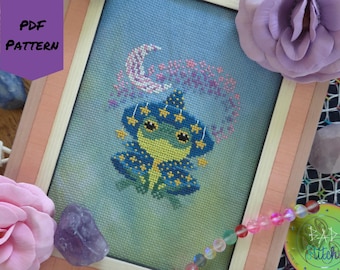 May include: Cross-stitch pattern of a whimsical frog wearing a star-studded hat and cape, framed in a light wood frame. The frog is green and yellow, with blue accents. The text "PDF PATTERN" is in the upper left corner.