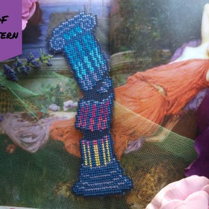 Op de afbeelding: Een blauwe, kruissteek bladwijzer met een geometrisch ontwerp, met tinten blauw, roze en geel. De bladwijzer wordt weergegeven op een pagina van een boek met een schilderij van een vrouw in een rode jurk. De woorden "PDF PATTERN" zijn zichtbaar.
