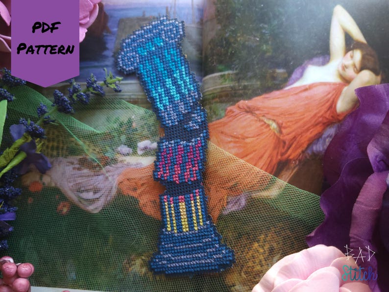 Op de afbeelding: Een blauwe, kruissteek bladwijzer met een geometrisch ontwerp, met tinten blauw, roze en geel. De bladwijzer wordt weergegeven op een pagina van een boek met een schilderij van een vrouw in een rode jurk. De woorden "PDF PATTERN" zijn zichtbaar.