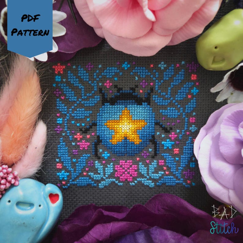 May include: A cross-stitch pattern featuring a blue and yellow star-shaped design within a square frame, surrounded by floral and leaf motifs in blue, pink, and purple. The words "PDF PATTERN" are in a blue box. The words "BAD Stitch" are in the bottom right corner.