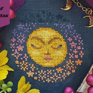 Puede incluir: Un patrón de punto de cruz con un sol amarillo con los ojos cerrados, rodeado por un anillo de estrellas rosas y naranjas, sobre un fondo azul oscuro. Las palabras "PDF PATTERN" están en una pancarta morada. Colgantes de luna dorados cuelgan de una cadena.