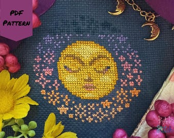 May include: A cross-stitch pattern featuring a yellow sun with closed eyes, surrounded by a ring of pink and orange stars, on a dark blue background. The words "PDF PATTERN" are in a purple banner. Gold moon charms hang from a chain.