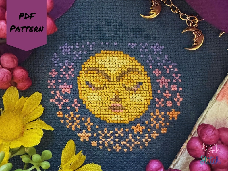 May include: A cross-stitch pattern featuring a yellow sun with closed eyes, surrounded by a ring of pink and orange stars, on a dark blue background. The words "PDF PATTERN" are in a purple banner. Gold moon charms hang from a chain.