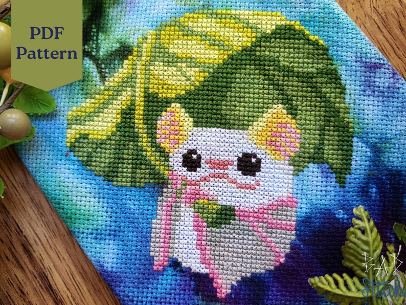 May include: Cross stitch pattern of a white animal with pink ears and a green leaf on its head. The animal is surrounded by a blue, green, and yellow background.