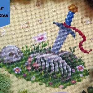 May include: Cross-stitch design showing a sword piercing a skeleton in a grassy scene, with small flowers and a skull. The sword has a blue handle and a red ribbon. The text "PDF PATTERN" is in the upper left corner.