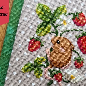 May include: Cross stitch pattern featuring a brown mouse sitting on a strawberry plant with green leaves, white flowers, and red strawberries. The pattern is on a beige fabric with white polka dots.